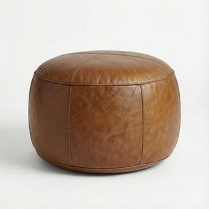 May include: A round, brown leather ottoman with a stitched top and vertical seams. The ottoman is a warm, medium brown color and has a smooth, slightly textured surface. It is a comfortable and stylish piece of furniture.