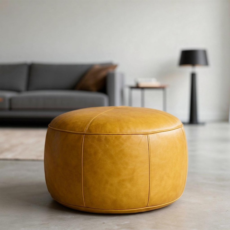 May include: A round, mustard-yellow leather ottoman. The ottoman has a stitched design and is placed on a concrete floor. A gray sofa and black floor lamp are visible in the background.