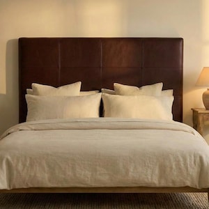 May include: A bed with a dark brown leather headboard and beige bedding. The bed is made with a wooden frame and has several pillows. A lamp sits on a small wooden side table.