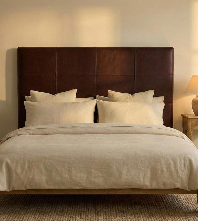 May include: A bed with a dark brown leather headboard and beige bedding. The bed is made with a wooden frame and has several pillows. A lamp sits on a small wooden side table.