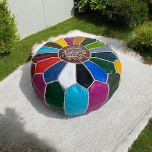 May include: A round, multi-colored leather pouf with a geometric design. The pouf features a variety of colors, including red, blue, green, and yellow, and is accented with white stitching. The top has a brown center with a white embroidered design.