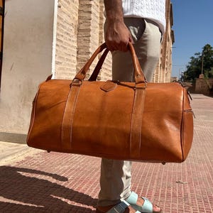 May include: A large, brown leather duffel bag with two top handles and a shoulder strap. The bag has a rectangular shape with a zipper closure and is held by a person. The bag is made of a smooth, textured leather.