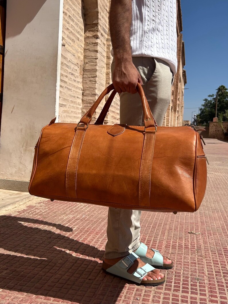 May include: A large, brown leather duffel bag with two top handles and a shoulder strap. The bag has a rectangular shape with a zipper closure and is held by a person. The bag is made of a smooth, textured leather.