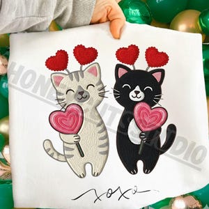 Könnte beinhalten: Weißer Stoff mit zwei Cartoon-Katzen, die herzförmige Lollis halten. Eine Katze ist grau und weiß, die andere schwarz und weiß. Beide Katzen tragen rote Herz-Stirnbänder. Das Wort "xoxo" ist unten geschrieben.