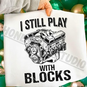 May include: White t-shirt featuring a detailed black and white illustration of an engine. The text "I STILL PLAY WITH BLOCKS" is printed above and below the engine graphic. The shirt is held up by a person.