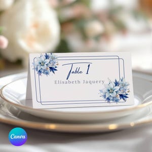Puede incluir: Tarjeta de mesa blanca con detalles florales azules y texto azul marino. La tarjeta dice "Table 1" y "Elisabeth Jaquery". La tarjeta está colocada sobre un plato blanco con borde dorado, con un fondo floral borroso.