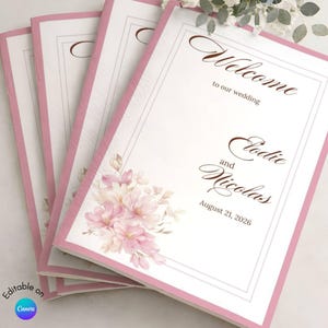 May include: A stack of rectangular wedding programs with a pink border and a white center. The text reads Welcome to our wedding, Elodie and Nicolas, and August 21, 2026. A cluster of light pink flowers is printed in the bottom left corner.