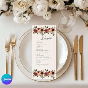 May include: A formal table setting with a menu card. The menu card is cream-colored with floral accents in burgundy, peach, and green. The menu lists a multi-course meal. Gold forks and knives are placed on either side of the plate.