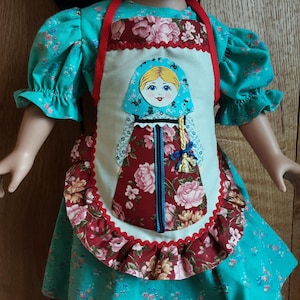 May include: A doll wearing a teal floral dress and a decorative apron. The apron features a Matryoshka doll design with a red floral skirt and a blue headscarf. The apron is trimmed with red and white ruffles.