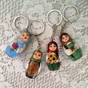 May include: Four painted wooden Russian nesting doll keychains. Each doll is wearing a different colorful dress and holding flowers. The keychains are attached to silver key rings.