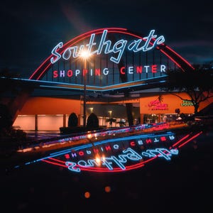 May include: A neon sign for Southgate Shopping Centre with red and blue lights. The sign is reflected in a wet surface.