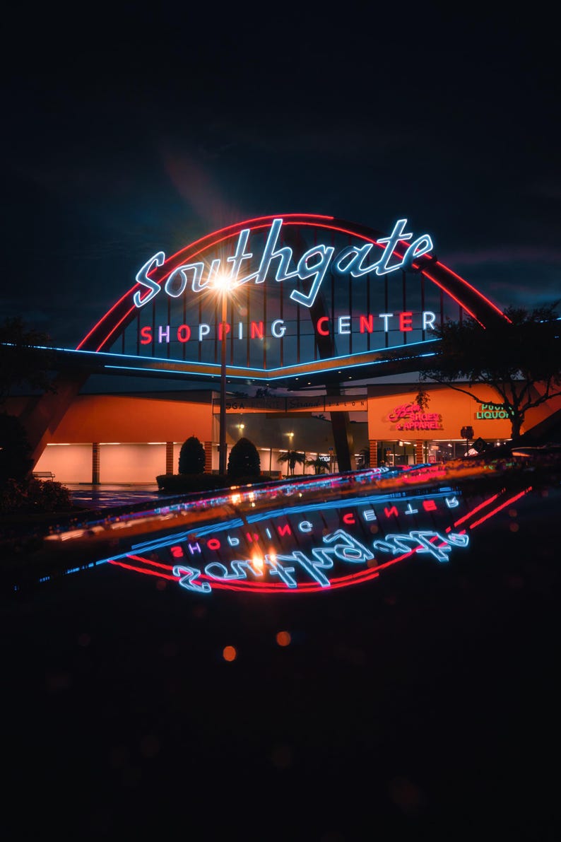 May include: A neon sign for Southgate Shopping Centre with red and blue lights. The sign is reflected in a wet surface.