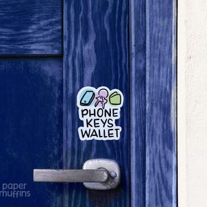 May include: A white sticker with the words "PHONE KEYS WALLET" and icons of a phone, keys, and a wallet. The sticker is affixed to a blue door with a silver handle. The background is a white wall.