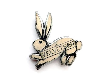 May include: A cream-colored rabbit pin with black outlines and detailing. The rabbit is depicted in a leaping pose with the word VELVETEEN printed in black capital letters across its midsection. The pin features a distressed, vintage-style finish.