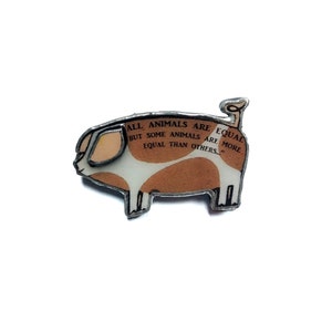 May include: A silver pig pin with a white and brown spotted pattern. The pig has a quote from George Orwell's Animal Farm printed on its side: "All animals are equal but some animals are more equal than others."
