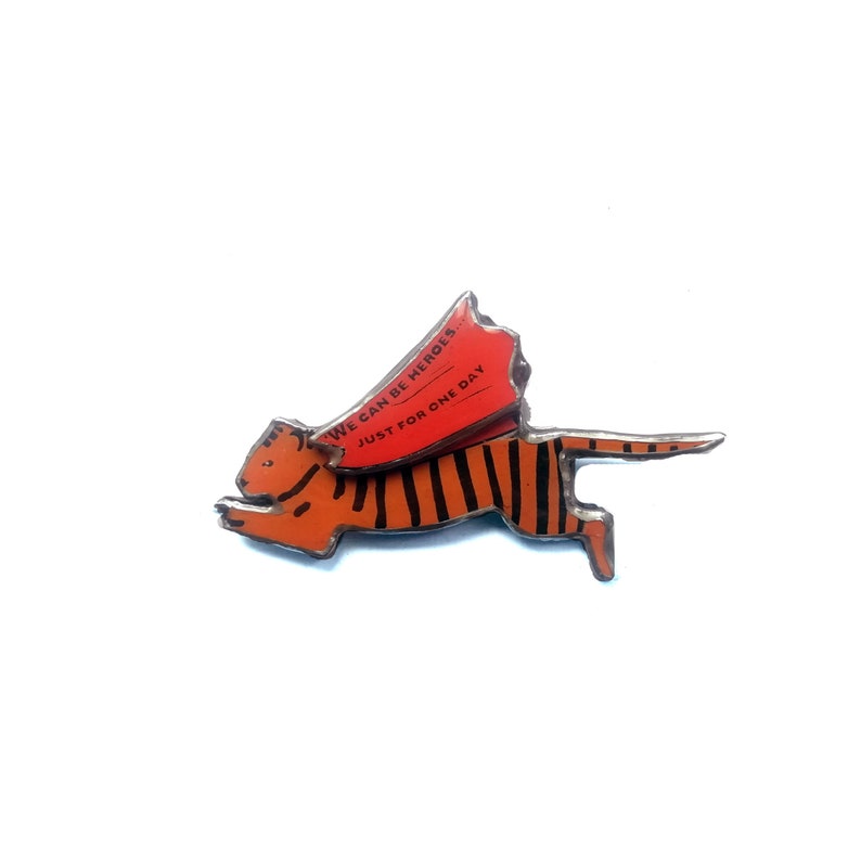 May include: A metal pin of a tiger wearing a red cape with the text "We can be heroes. Just for one day."