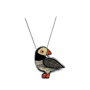 May include: A silver-toned necklace featuring a pendant shaped like a puffin. The bird design incorporates black, white, and orange colors with printed text on the body that reads A PERFECT PUFFIN FAVOURITE. The pendant has a dark metal border.