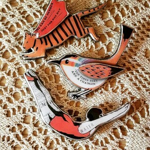May include: Three metal pins with colorful illustrations and text. The first pin features a tiger with the text "We can be heroes... Just for one day". The second pin features a bird with the text "The times they are a changin'". The third pin features a diver with the text "Water".