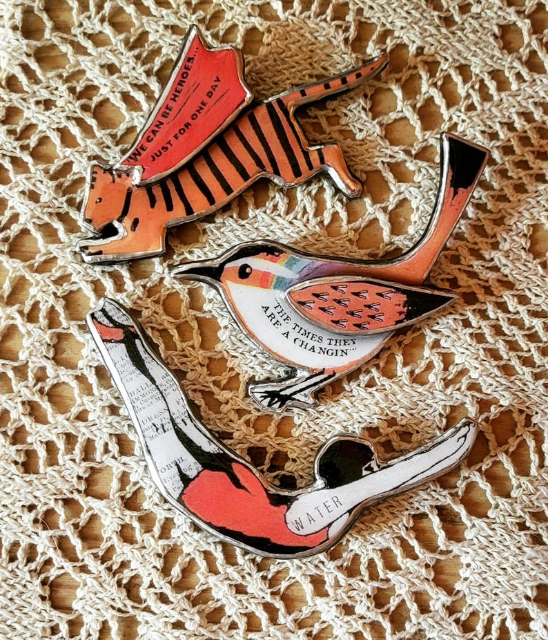 May include: Three metal pins with colorful illustrations and text. The first pin features a tiger with the text "We can be heroes... Just for one day". The second pin features a bird with the text "The times they are a changin'". The third pin features a diver with the text "Water".