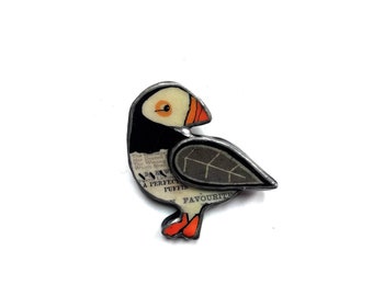 May include: A handcrafted puffin bird brooch with a vintage aesthetic. The bird features a black and white body, a cream-colored face, and an orange beak and feet. The brooch incorporates text from a vintage book and is outlined in silver.