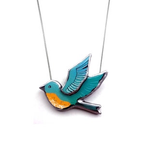 Può includere: Una collana con un ciondolo a forma di uccello blu turchese con accenti gialli. L'uccello sta volando con le ali spiegate. La collana è in metallo e ha una catena argentata.