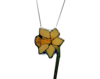 Può includere: Una collana con un ciondolo a forma di narciso giallo su una catena d'argento. Il fiore è dettagliato con un centro giallo e uno stelo verde.