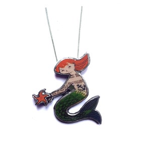 May include: A silver necklace with a mermaid charm. The mermaid is made of metal and has a green tail, red hair, and a printed design on her torso. She is holding a starfish in her hand.