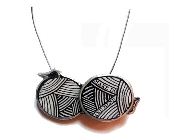 Puede incluir: Un collar de plata con dos ovillos de lana negros y blancos como colgantes. Los ovillos de lana tienen el texto "Make Do and Mend" impreso en ellos.
