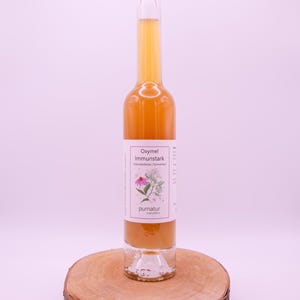 Könnte beinhalten: Eine klare Glasflasche gefüllt mit einer goldenen Flüssigkeit, verschlossen mit einem Korken. Die Flasche hat ein weißes Etikett mit Text und einer floralen Illustration. Sie steht auf einer runden Holzscheibe, vor einem zartrosa Hintergrund. Gesundheitsprodukt.