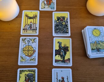 Può includere: Un layout di carte dei tarocchi con varie carte illustrate, tra cui una con un cavaliere a cavallo. Due candele bianche affiancano la disposizione su una superficie di legno. Le carte sono disposte secondo uno schema specifico.