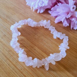 Könnte beinhalten: Ein Armband aus Rosenquarz-Chips. Es besteht aus kleinen, unregelmäßig geformten, hellrosa Steinen. Der Hintergrund zeigt zarte rosa Blüten und eine Holzoberfläche. Das Armband ist ca. 18 cm lang.