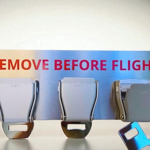 May include: A metal sign with the words "REMOVE BEFORE FLIGHT" in red lettering. Below the sign are three silver airplane seat belt buckles. One buckle is unfastened, with the belt detached.