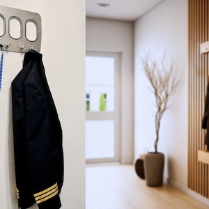 May include: A silver metal wall-mounted coat rack with three oval openings and four hooks. A black jacket with gold stripes hangs from the hooks, along with a blue lanyard. The rack is mounted on a white wall.