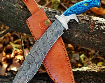 May include: A Damascus steel hunting knife with a blue wooden handle and a brown leather sheath. The blade has a unique swirling pattern. The sheath is secured with a leather strap and a metal button.