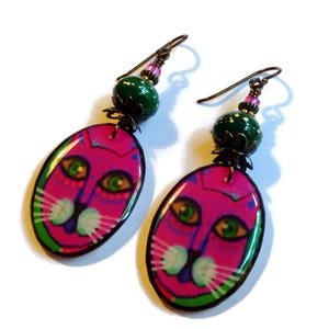 May include: A pair of pink cat face earrings with green and white accents. The earrings have a green bead and a bronze charm.
