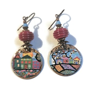 May include: A pair of earrings with a red bead and a round copper disc with a colorful painted scene of houses and a tree.