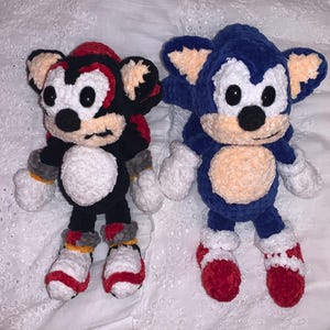 May include: Two handmade crochet plush toys: one black and red, the other blue. The black and red plush has a white chest and red accents. The blue plush has a tan chest and red shoes. Both have black eyes and tan ears.
