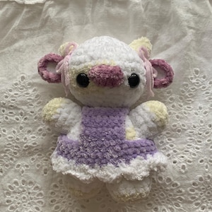 May include: A handmade crochet cow plushie with a white body, a purple dress, and black eyes. The cow has pink and yellow accents on its ears and snout. The plushie is soft and cuddly, perfect for a child's toy or nursery decor.