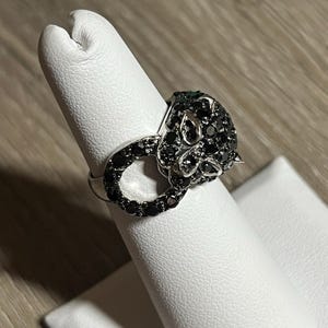 May include: A silver ring with a panther head design, embellished with black gemstones. The ring is displayed on a white stand, highlighting its intricate details. The panther's eyes are green. A stylish piece of jewellery.