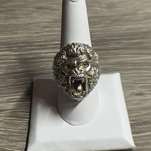 Puede incluir: Un anillo de plata con un diseño detallado de cabeza de león. La boca del león está abierta y sus dientes son visibles. El anillo está en un expositor de anillos blanco.