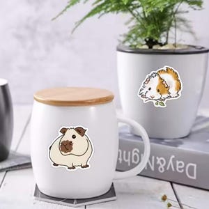 Peut inclure: Mug en c&eacute;ramique blanche avec un couvercle en bois, orn&eacute; d'un autocollant de cochon d'Inde. Un deuxi&egrave;me autocollant de cochon d'Inde est sur un pot blanc avec une plante. Les autocollants sont beige, marron et blanc.