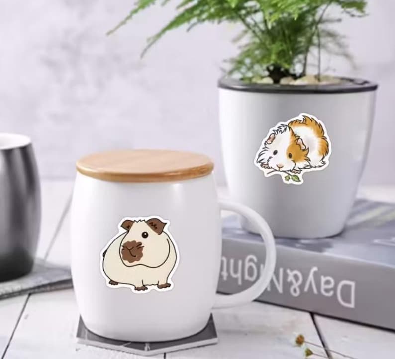 Peut inclure: Mug en c&eacute;ramique blanche avec un couvercle en bois, orn&eacute; d'un autocollant de cochon d'Inde. Un deuxi&egrave;me autocollant de cochon d'Inde est sur un pot blanc avec une plante. Les autocollants sont beige, marron et blanc.