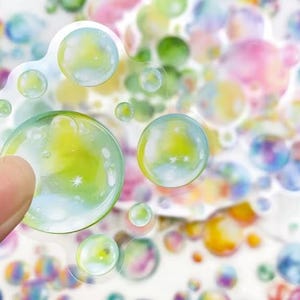 May include: A selection of bubble stickers with a transparent, glossy finish. The stickers display a range of sizes and colours, including green, yellow, blue, and pink. One sticker is held by a finger, highlighting the sticker's design.