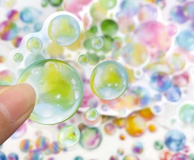 May include: A selection of bubble stickers with a transparent, glossy finish. The stickers display a range of sizes and colours, including green, yellow, blue, and pink. One sticker is held by a finger, highlighting the sticker's design.
