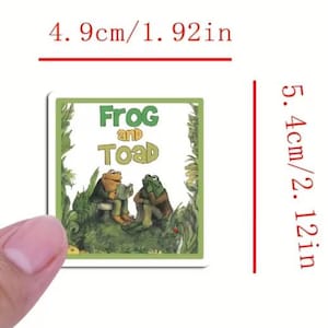 以下が含まれることがあります： 「Frog and Toad」の表紙が描かれた正方形のステッカー。ステッカーの幅は4.9cm、高さは5.4cmです。緑と茶色の背景に、2匹の人格化されたカエルが描かれており、タイトルテキストは緑色です。