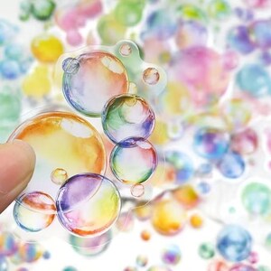 May include: A selection of bubble stickers in different sizes and colours. The stickers have a watercolour effect with shades of yellow, orange, green, blue, and purple. Some stickers are held by a hand, while others are scattered in the background.