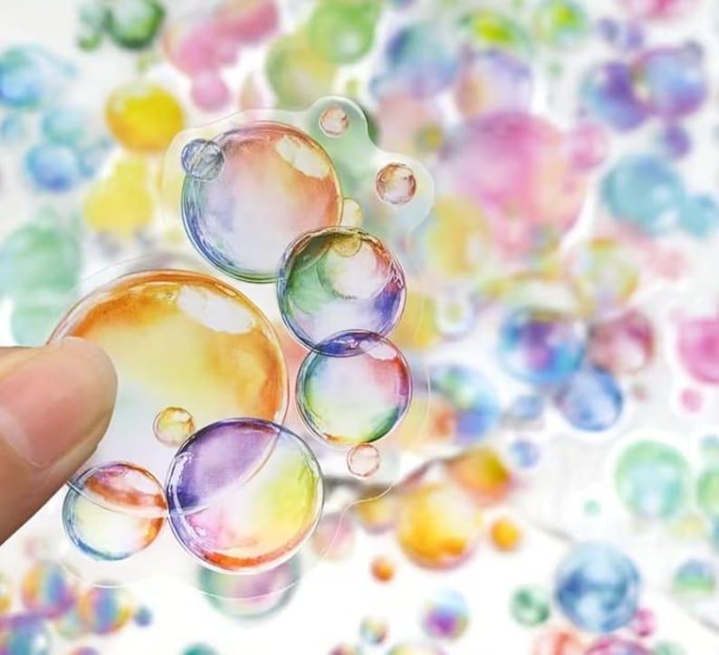 May include: A selection of bubble stickers in different sizes and colours. The stickers have a watercolour effect with shades of yellow, orange, green, blue, and purple. Some stickers are held by a hand, while others are scattered in the background.