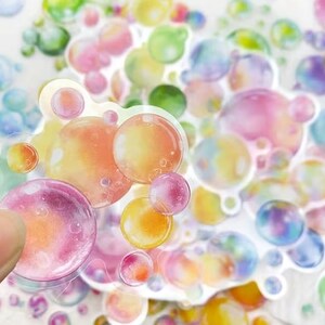 May include: A selection of bubble stickers in a range of colours, including pink, orange, yellow, green, and blue. The stickers vary in size and are on a white background. One sticker is being held by a finger. The stickers have a glossy finish.