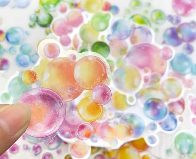 May include: A selection of bubble stickers in a range of colours, including pink, orange, yellow, green, and blue. The stickers vary in size and are on a white background. One sticker is being held by a finger. The stickers have a glossy finish.
