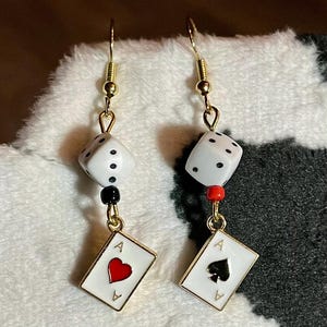 May include: A pair of gold earrings with a white dice charm and a playing card charm. One earring has a red ace of hearts charm and the other has a black ace of spades charm.